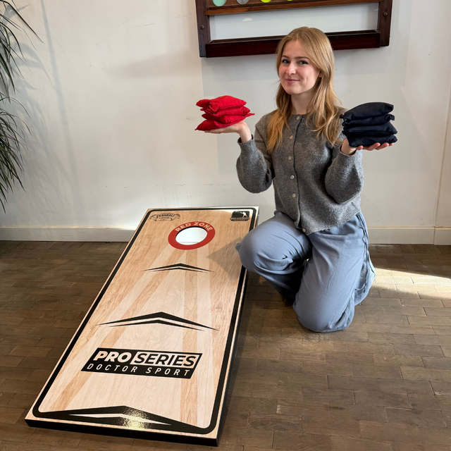 Eine lächelnde junge Frau kniet neben einem Cornhole-Brett der Pro Series und hält rote und blaue Wurfsäcke in den Händen.