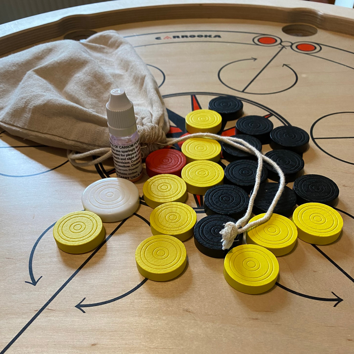 Carrooka Carrom360 Spielsteine in Gelb, Schwarz und Rot, weißer Striker, Fläschchen Startpulver und Baumwollbeutel mit Kordelzug