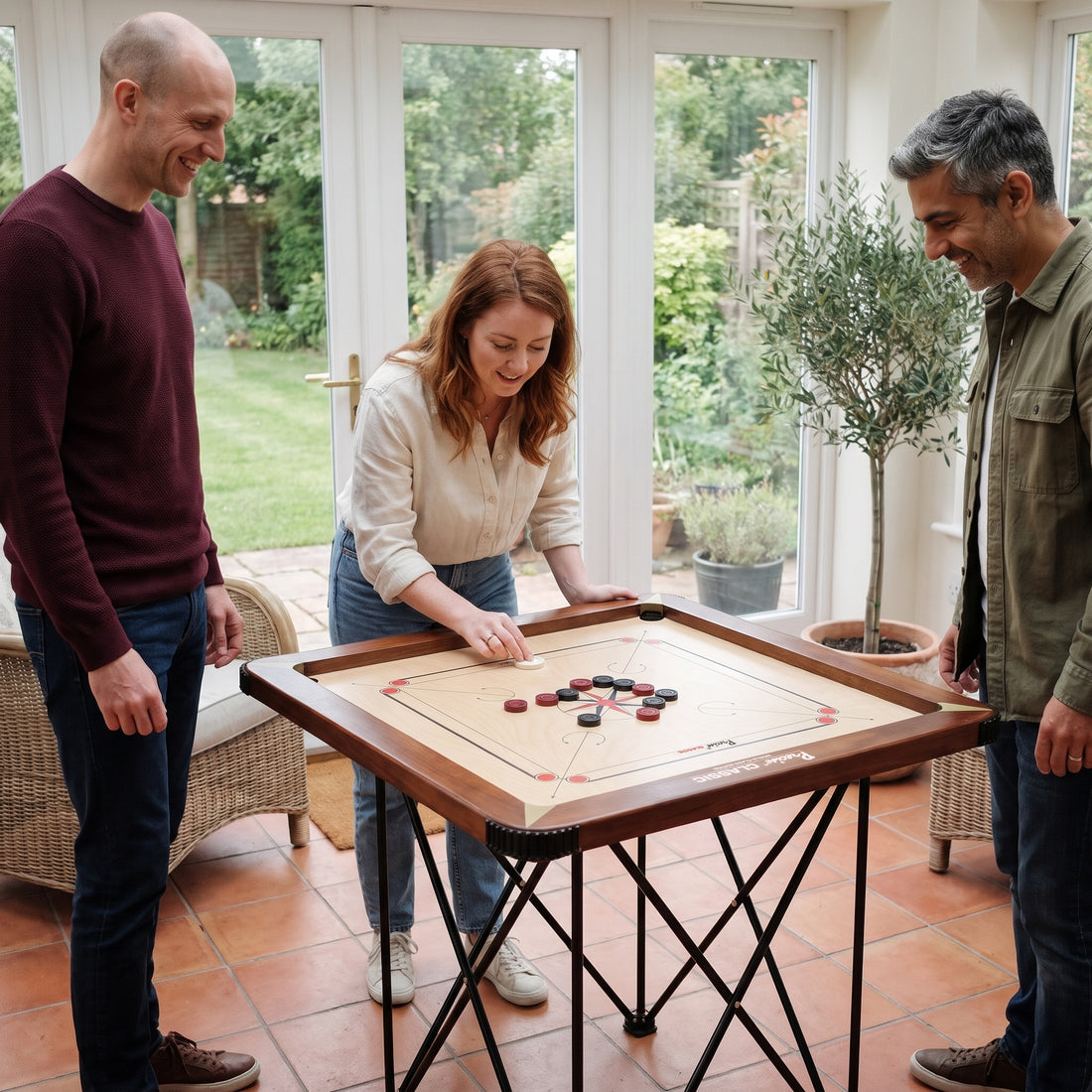 Precise Carrom Champion 24mm World-Class Edition auf dem klappbaren Carrom-Tisch, drei Freunde spielen Carrom im Wintergarten