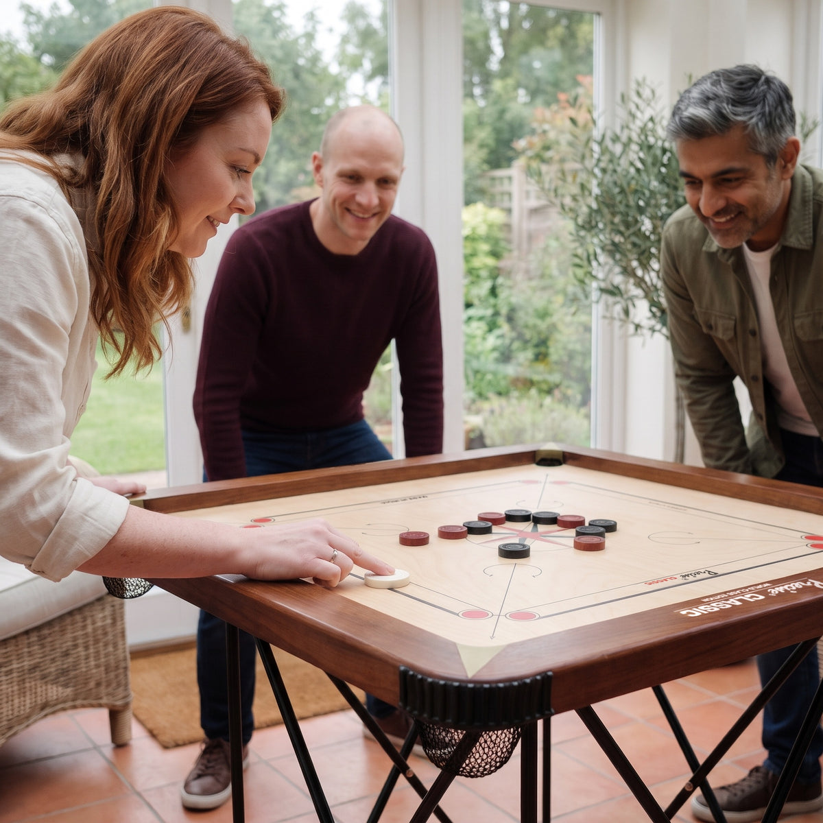 Precise Carrom Champion 24mm World-Class Edition im Wintergarten: eine Spielerin schnippt den Striker, zwei Mitspieler schauen gespannt zu