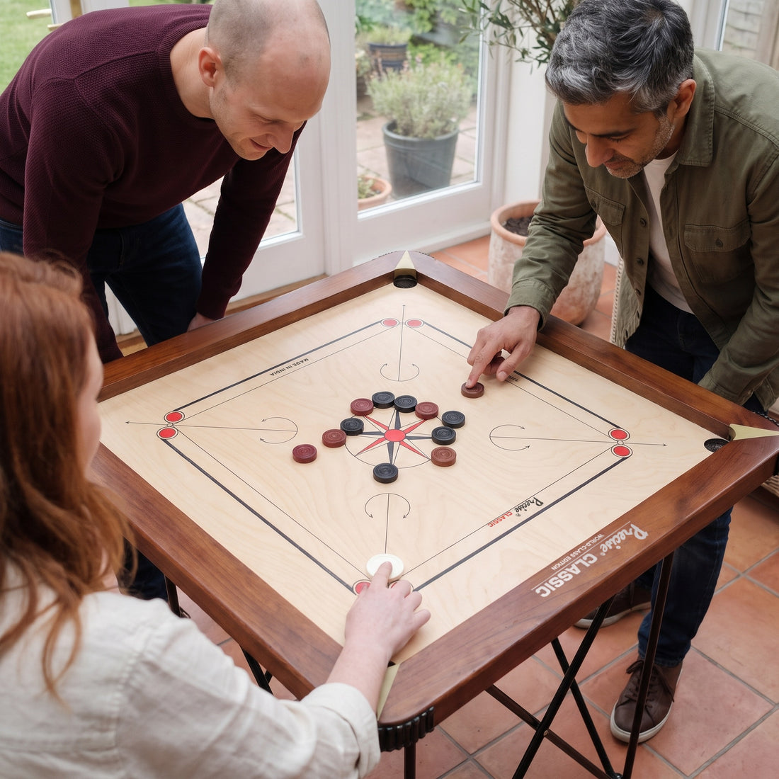 Precise Carrom Champion 24mm World-Class Edition aus der Nähe: drei Freunde spielen im Wintergarten, Brett mit walnussfarbenem Rahmen und Spielsteinen gut sichtbar